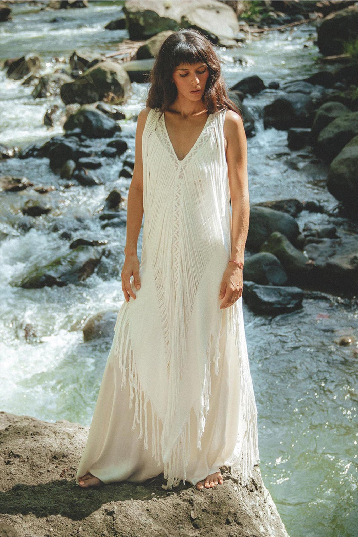 Standing on a rock by a flowing stream, the individual dons the See Through Dress, a long maxi dress with exquisite handmade embroidery by AYA Sacred Wear. The lush greenery enhances the serene beauty of this tribal raw cotton cover-up.