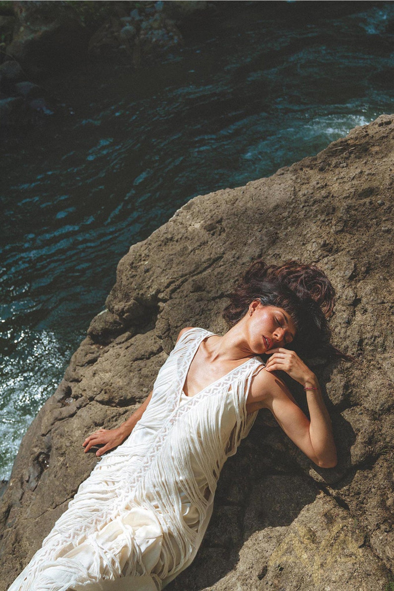 Dressed in AYA Sacred Wear's See Through Dress • Long Maxi Dress • Tribal Raw Cotton Cover-up, a woman reclines on rocks by the river. Sunlight dances across the textured fabric, while the nearby flowing water shimmers. Her dark hair cascades around her, and she exudes relaxation and serenity.