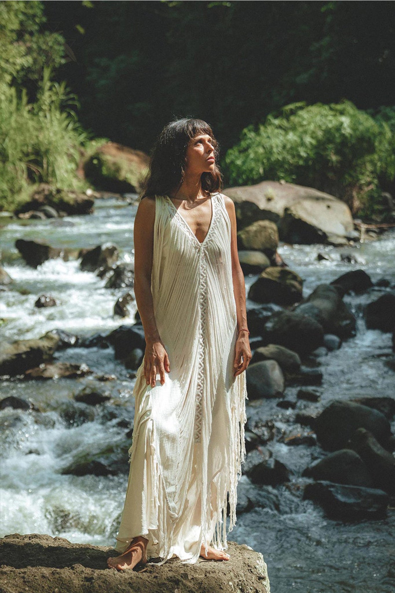 Standing atop a rock by a flowing river, an individual is elegantly dressed in the AYA Sacred Wear's See Through Dress – a Long Maxi Dress in Tribal Raw Cotton. The intricate handmade embroidery adds charm to the sleeveless white cover-up, set against a backdrop of lush greenery illuminated by natural daylight.