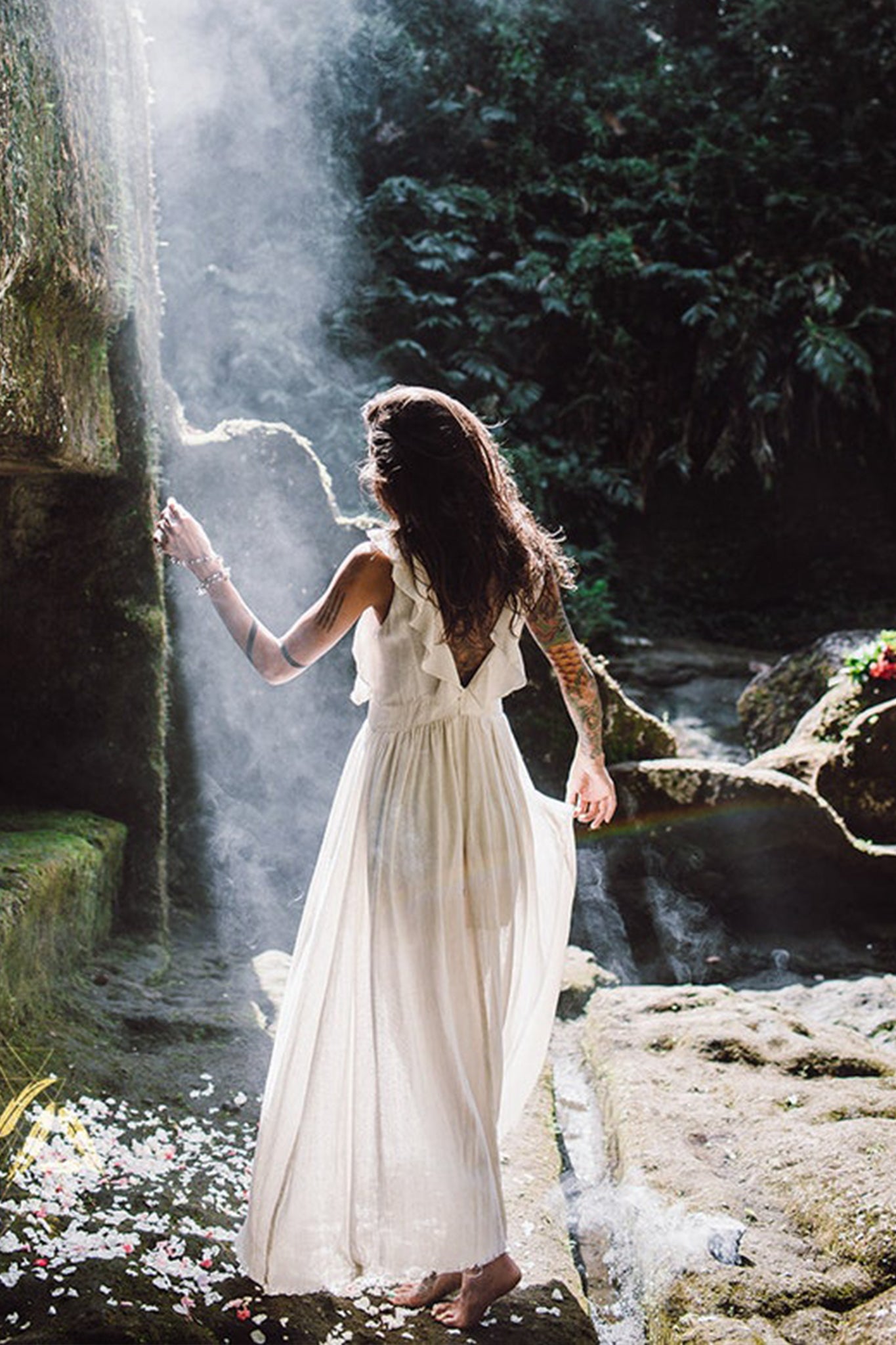 Wearing the Simple Boho Bohemian Wedding Dress by AYA Sacred Wear, a woman stands on a rocky terrain amidst vibrant greenery, with gentle sunlight streaming through. She faces away, her hand resting on a rock wall, while petals are strewn across the ground.