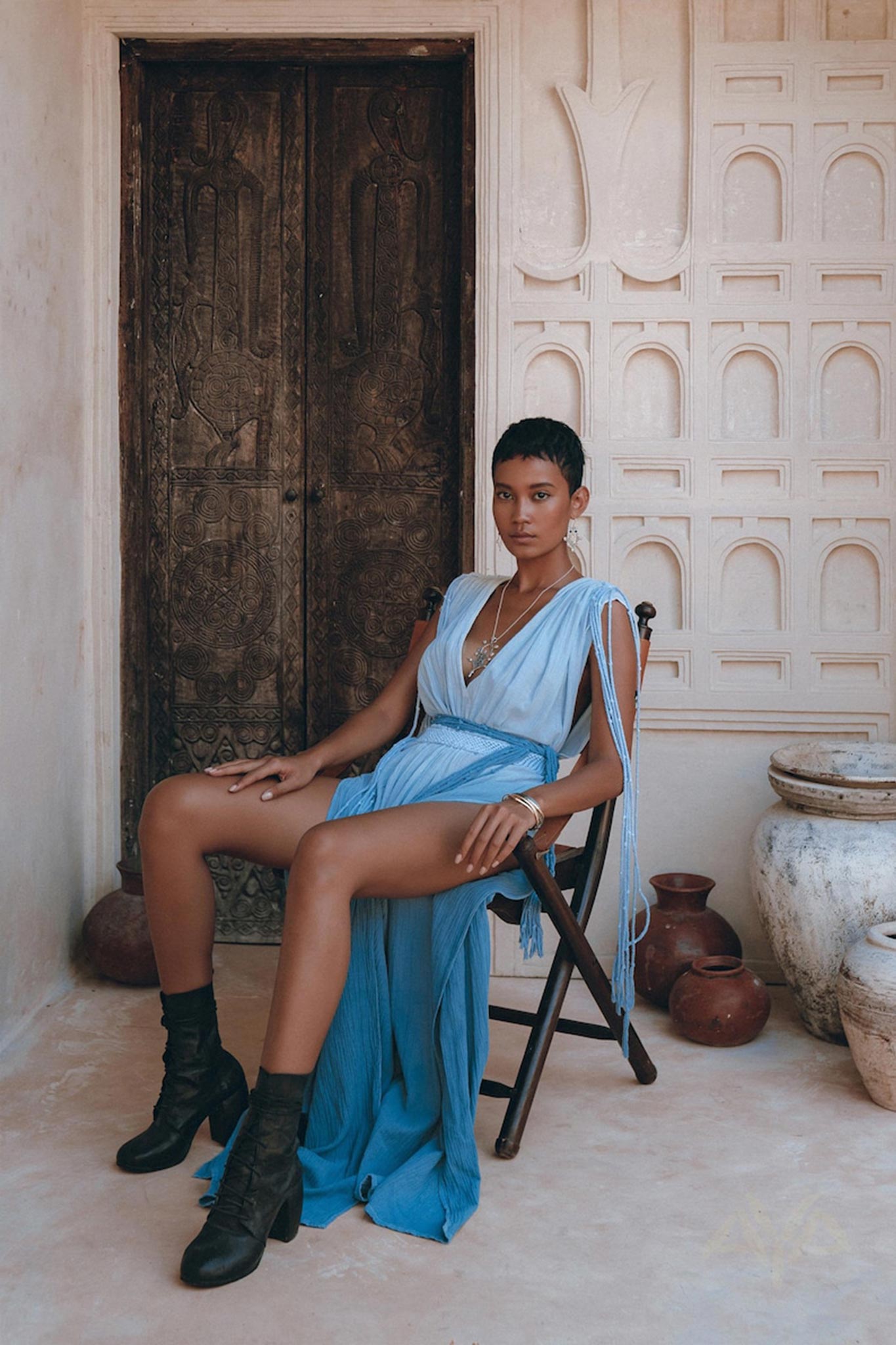 A person wearing a Sky Blue Ombre Dyed Dress from AYA Sacred Wear, complemented by black boots, sits on a wooden chair in front of an intricately carved wooden door. The scene is enhanced by pottery and textured beige walls with ornate architectural details.