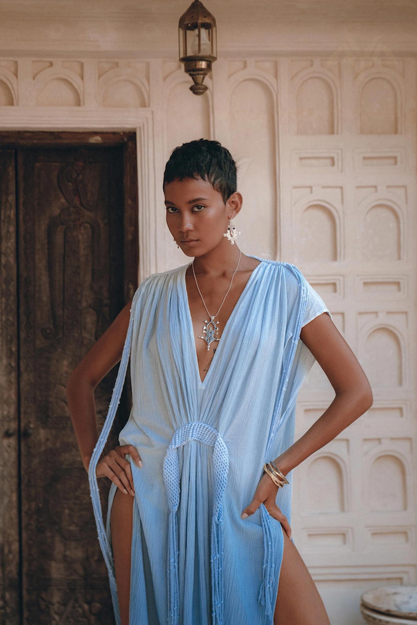 A person poses confidently in an AYA Sacred Wear creation, the Sky Blue Ombre Dyed Dress. This handcrafted boho wedding dress, complete with a deep V-neckline and high slits, elegantly boasts flowing fabric. They accessorize with a necklace and earrings, set against the backdrop of a decorative wall and an ornate wooden door.