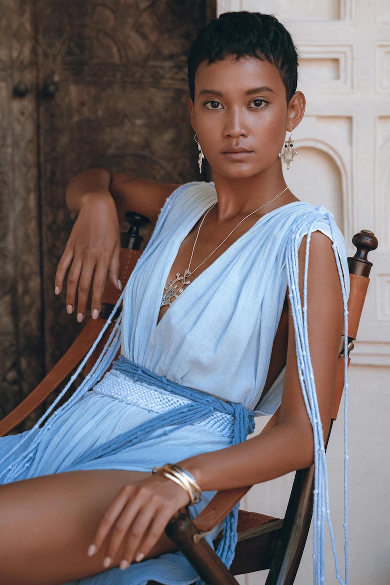 A person with short hair sits confidently in a wooden chair, adorned in an AYA Sacred Wear Sky Blue Ombre Dyed Dress, a flowing, sleeveless boho wedding dress complemented by silver jewelry. The handcrafted macrame design elegantly drapes across the ornate, textured background.