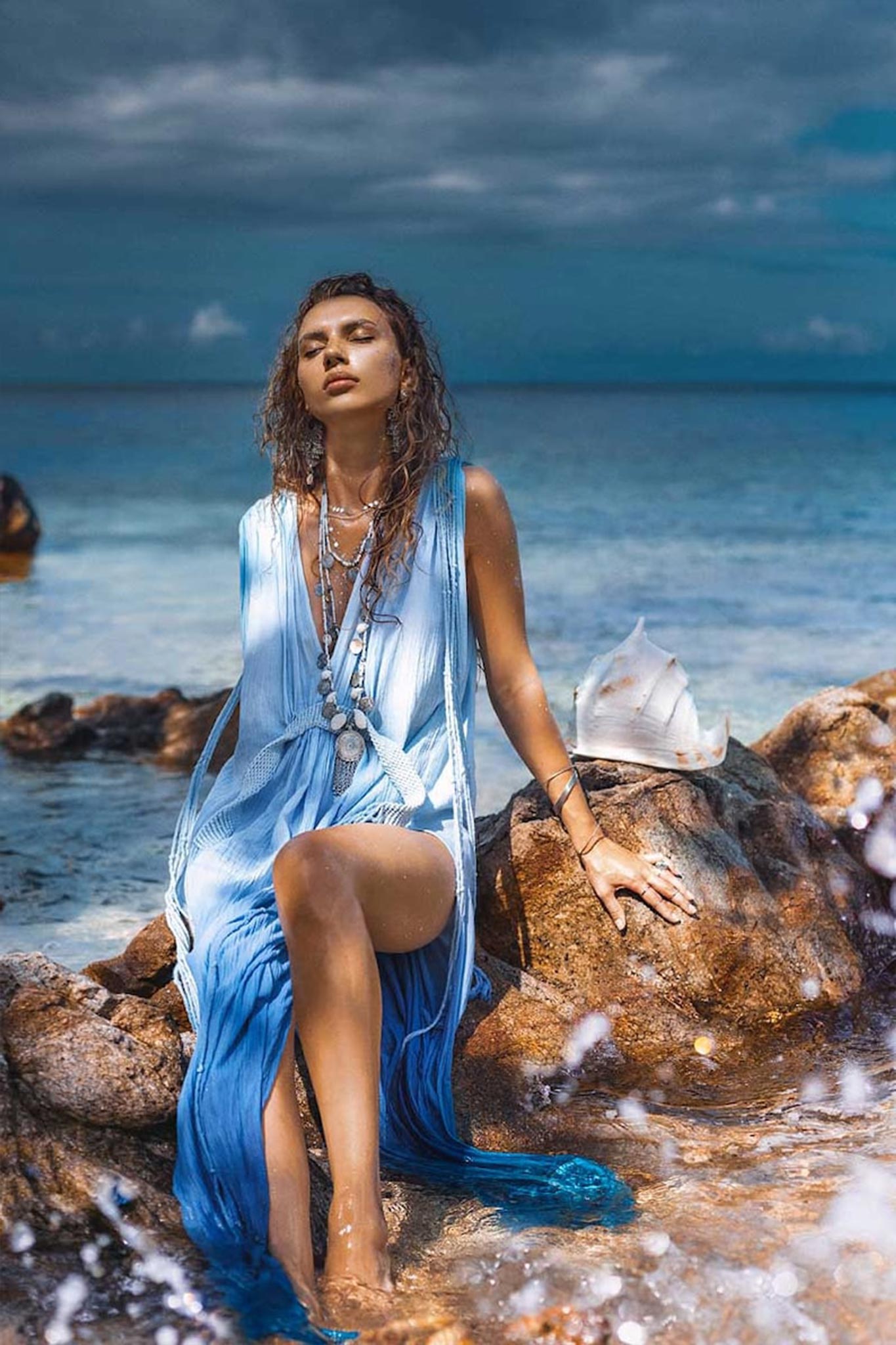 A woman in a Sky Blue Ombre Dyed Dress by AYA Sacred Wear sits on rocks by the ocean. Her curly hair cascades over layered necklaces, with a large conch shell beside her. The sky is overcast, and waves splash nearby.