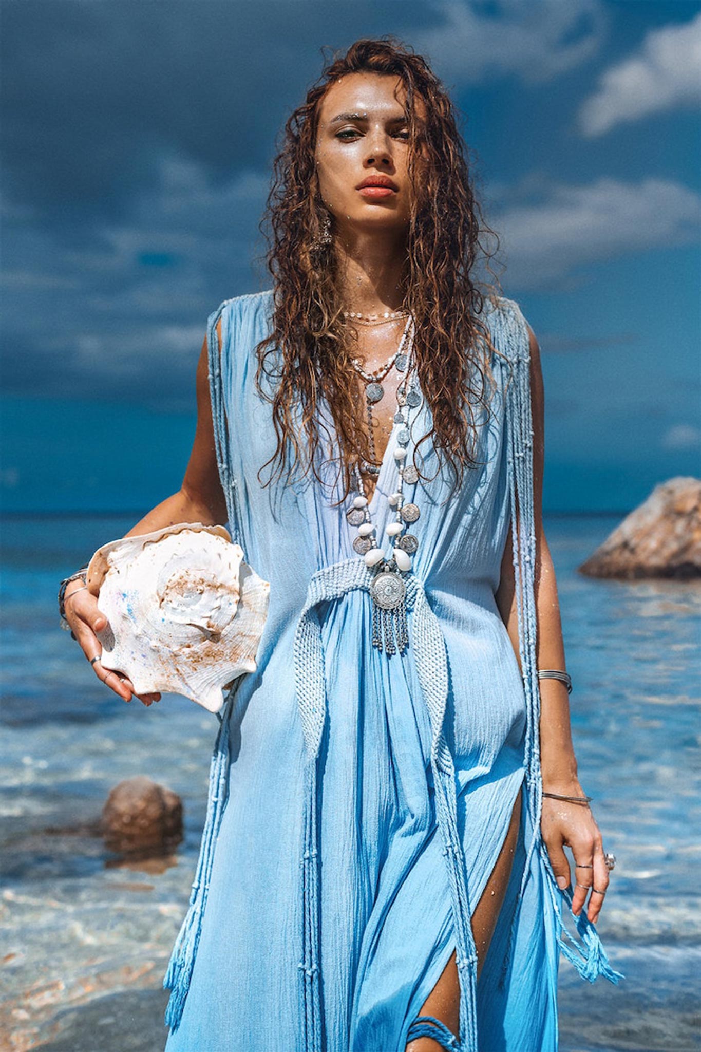 A woman stands on a beach holding a large seashell. Her long wet hair cascades over an AYA Sacred Wear Sky Blue Ombre Dyed Dress, a boho wedding dress adorned with handmade macrame details. The blue fabric of the dress mirrors the ocean's shades, its flowing design harmonizing beautifully with the serene backdrop of ocean and rocks under the cloudy sky.