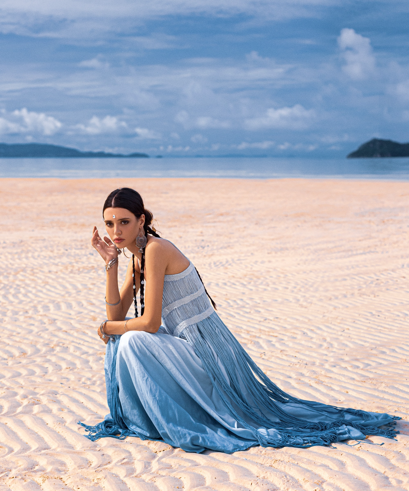 A woman sits on a sunlit beach in an AYA Sacred Wear Sky Blue Greek Goddess Dress, crafted from botanically dyed organic cotton. Her braided hair complements the partly cloudy sky and distant islands in the background as she gazes at the camera.