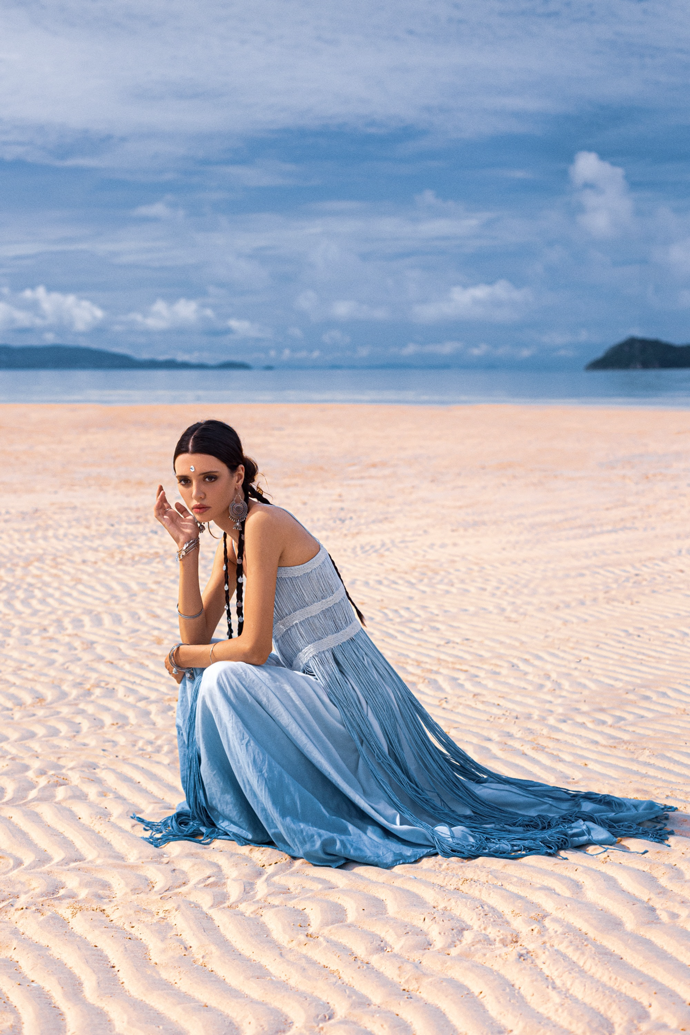 A woman sits on a sunlit beach in an AYA Sacred Wear Sky Blue Greek Goddess Dress, crafted from botanically dyed organic cotton. Her braided hair complements the partly cloudy sky and distant islands in the background as she gazes at the camera.