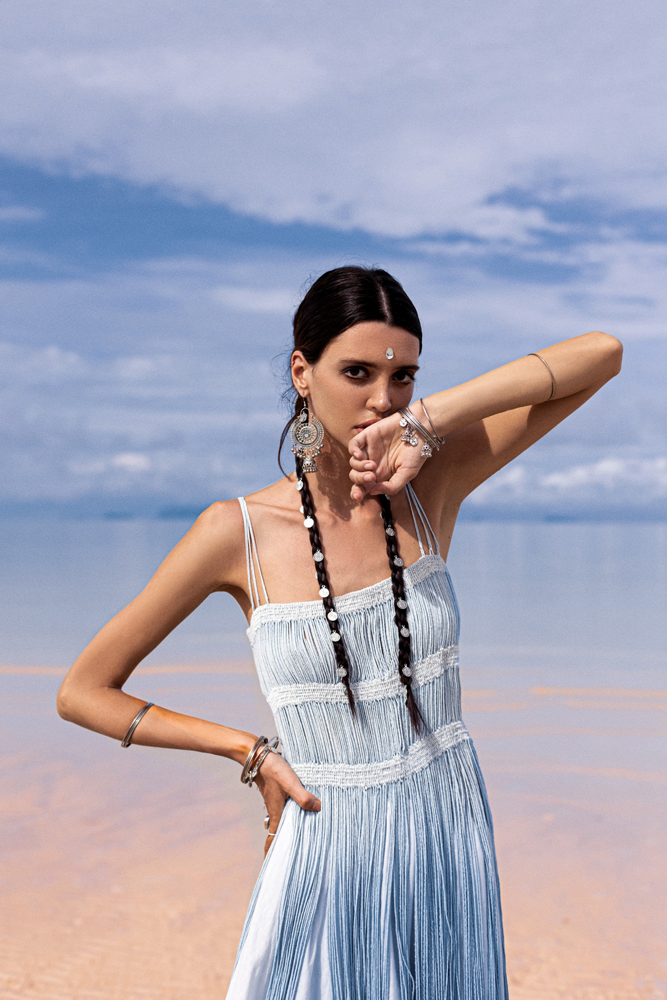 On the beach, a woman in a Sky Blue Greek Goddess Dress by AYA Sacred Wear stands with two braided hair adorned with beads. Her arm showcases silver bracelets against the peaceful beach and cloudy sky.