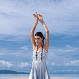 A woman in an AYA Sacred Wear Sky Blue Greek Goddess Dress stands on a beach with raised arms. The partly cloudy sky and ocean backdrop highlight her tribal-themed outfit, accented by long earrings and bracelets catching the coastal breeze.
