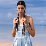A woman with long braided hair stands on a sandy beach wearing AYA Sacred Wear's Sky Blue Greek Goddess Dress, sleeveless and crafted from organic cotton. Intricate earrings and bracelets enhance her look as the dress's botanically dyed fabric complements the sea and sky behind her cloudy backdrop.