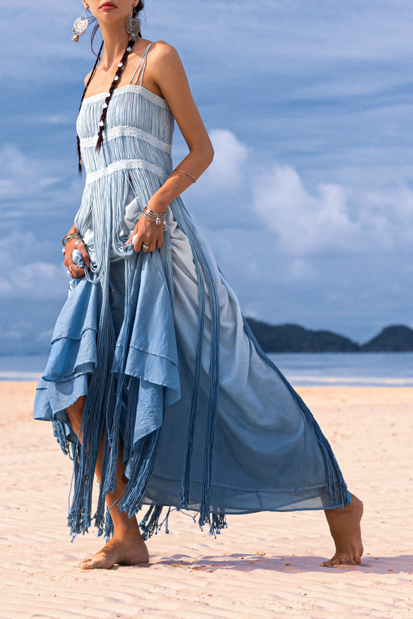 Barefoot on a sandy beach, a person wears AYA Sacred Wear's Sky Blue Greek Goddess Dress, an ombre macrame piece with spaghetti straps and botanical dyes. It complements their long braids and large earrings under a partly cloudy sky with distant hills visible.