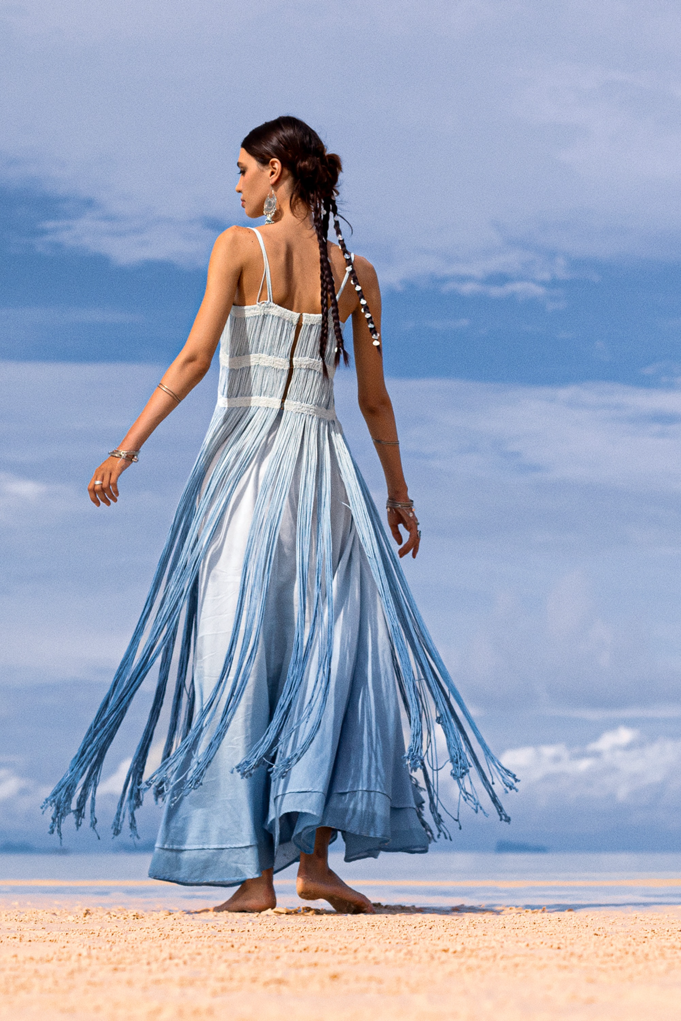 Barefoot on sand, a woman with braided hair wears the Sky Blue Greek Goddess Dress from AYA Sacred Wear. Made of botanically dyed organic cotton with long fringe, the dress elegantly flows as she gazes at a partly cloudy horizon.