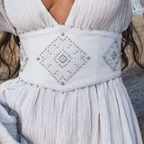 A woman wears the Tatva Dress in Celestial Mist Blue by AYA Sacred Wear, featuring a textured fabric, deep V-neck, and a wide handcrafted belt with geometric diamond patterns. The light airy dress complements her loosely flowing hair.