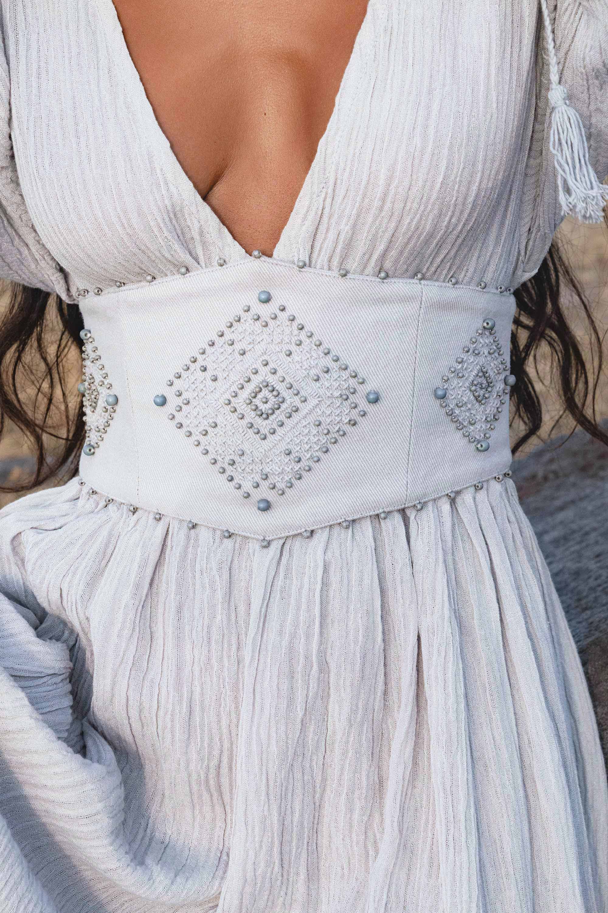 A woman wears the Tatva Dress in Celestial Mist Blue by AYA Sacred Wear, featuring a textured fabric, deep V-neck, and a wide handcrafted belt with geometric diamond patterns. The light airy dress complements her loosely flowing hair.