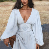 A woman with long, wavy dark hair stands outdoors on a rock wearing the AYA Sacred Wear Tatva Dress in Celestial Mist Blue—a botanically dyed dress with a handcrafted belt and deep V-neck, set against dry grass, green bushes, and trees.