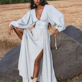 A woman poses outdoors in the AYA Sacred Wear Tatva Dress in Celestial Mist Blue, featuring billowy sleeves and a high slit. She styles it with white boots, a handcrafted belt, and lets her long wavy hair flow against trees and dry grass.