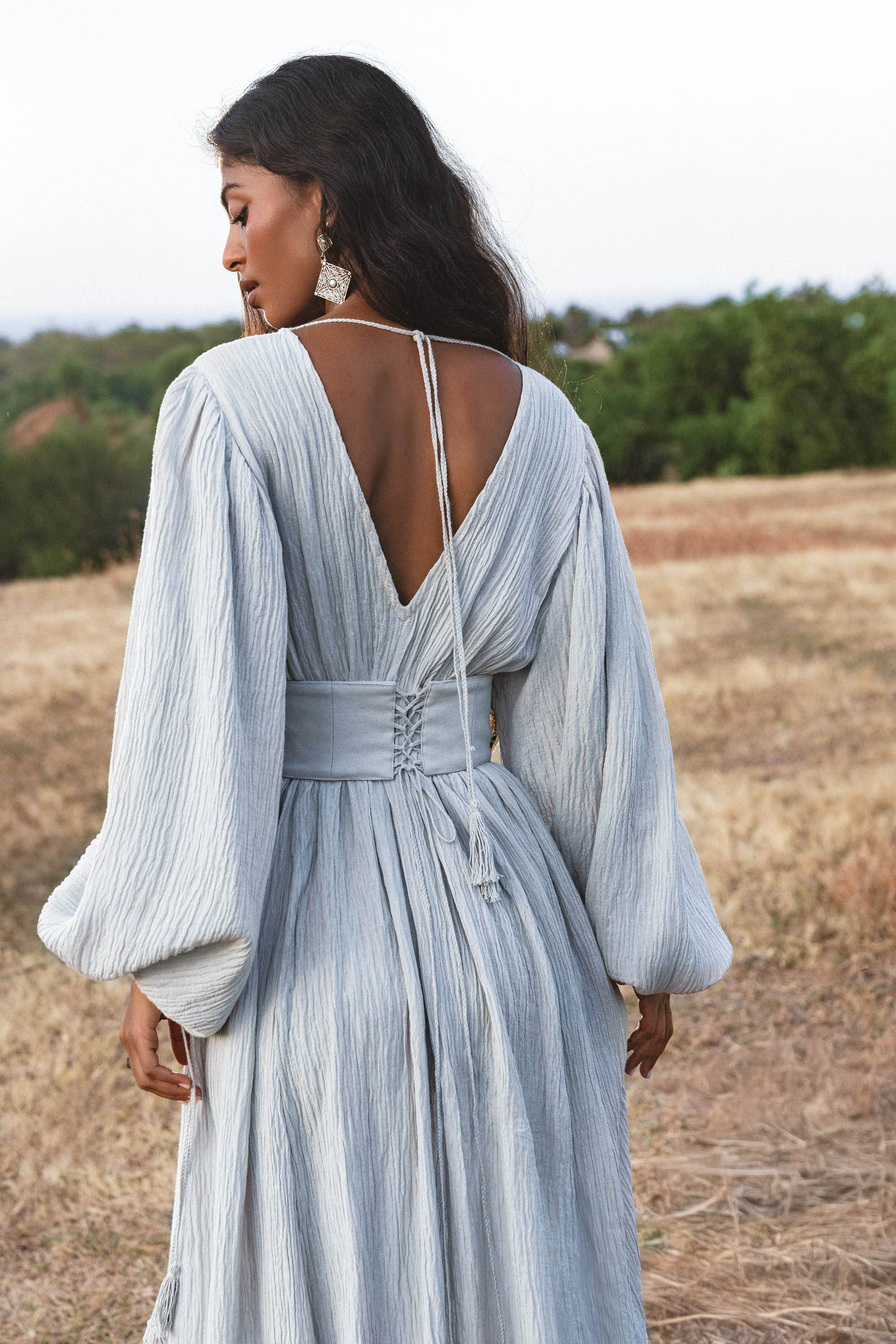 A woman stands in a field wearing the Tatva Dress Celestial Mist Blue by AYA Sacred Wear, featuring botanical dye, a deep V-back, wide sleeves, and a handcrafted belt. She faces away, highlighting her flowing hair and bold earrings.