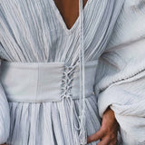 Close-up of a person wearing the AYA Sacred Wear Tatva Dress in Celestial Mist Blue, featuring botanical dye, textured fabric, a deep V-neck, puffy sleeves, and a handcrafted lace-up belt with tassel; hands rest on hips.