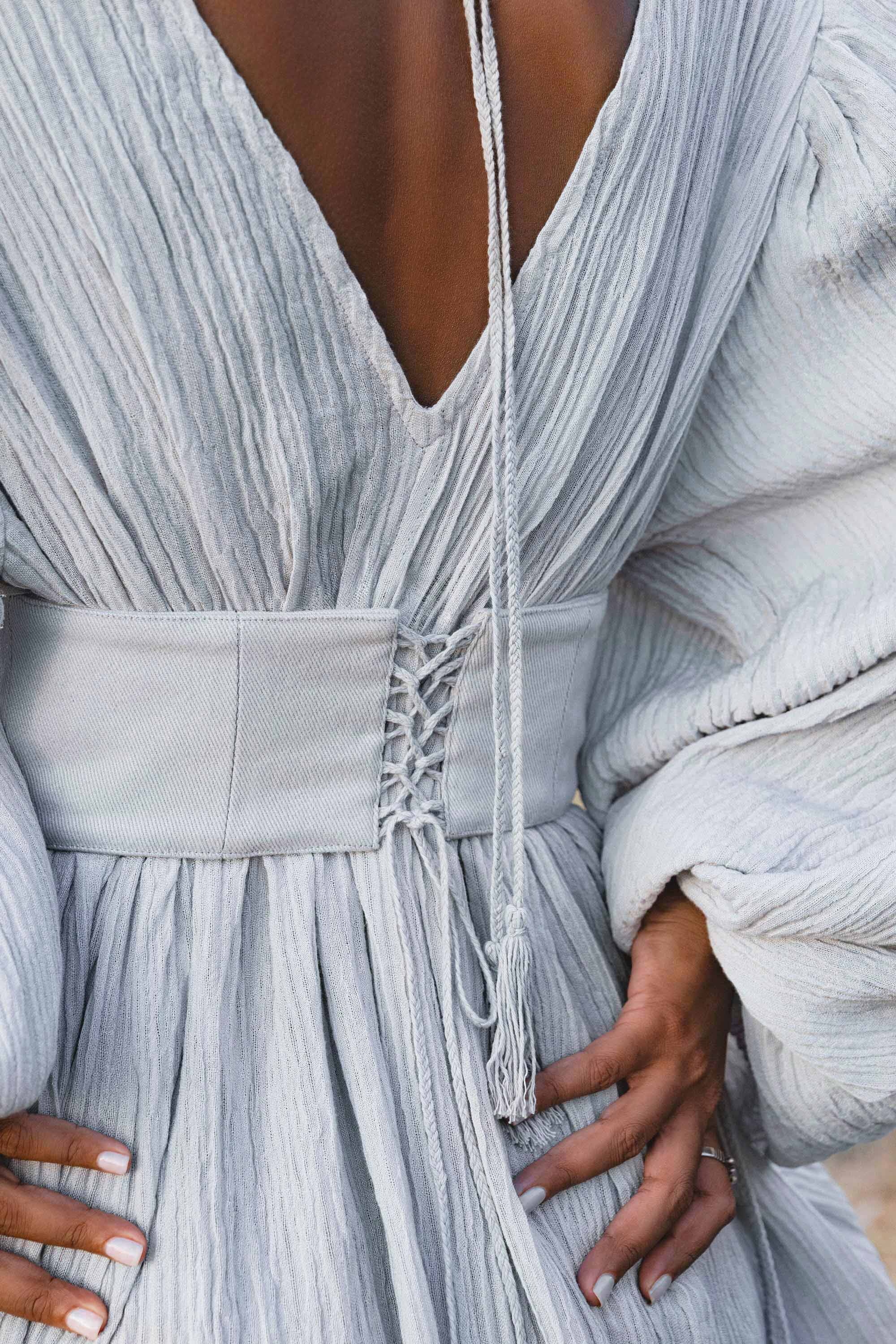 Close-up of a person wearing the AYA Sacred Wear Tatva Dress in Celestial Mist Blue, featuring botanical dye, textured fabric, a deep V-neck, puffy sleeves, and a handcrafted lace-up belt with tassel; hands rest on hips.