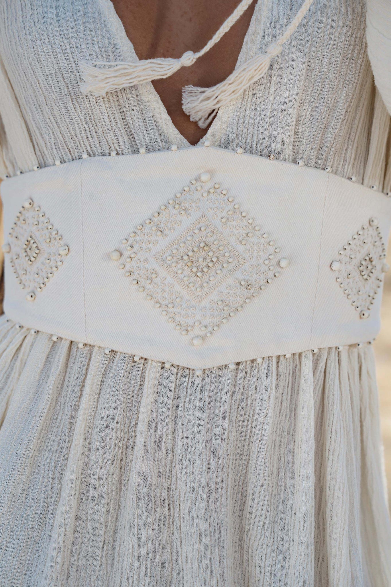 A close-up of the AYA Sacred Wear Tatva Dress Ivory Colour – Sustainable Bohemian Dress highlights its white botanically dyed fabric, wide handcrafted belt with pearl-beaded diamond motifs, and tasseled V-neck tie.