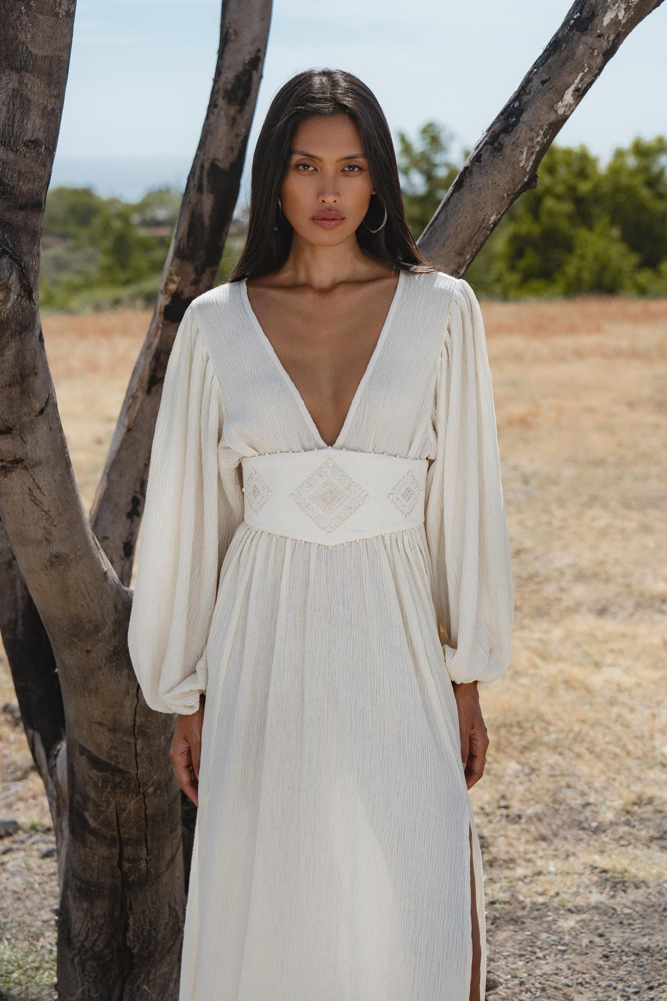 A woman with long dark hair stands outdoors by a tree, wearing the Tatva Dress Ivory Colour—a sustainable, bohemian dress by AYA Sacred Wear with long sleeves, a deep V-neck, and a wide handcrafted embroidered belt. Nature surrounds her.