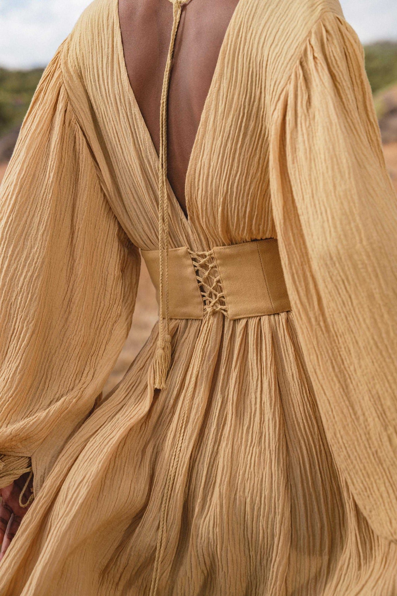 A person wears the Tatva Dress in Saffron Gold by AYA Sacred Wear—textured, pleated, with voluminous sleeves, a deep V-back, and a handcrafted lace-up belt. The outdoor setting is softly blurred in the background.