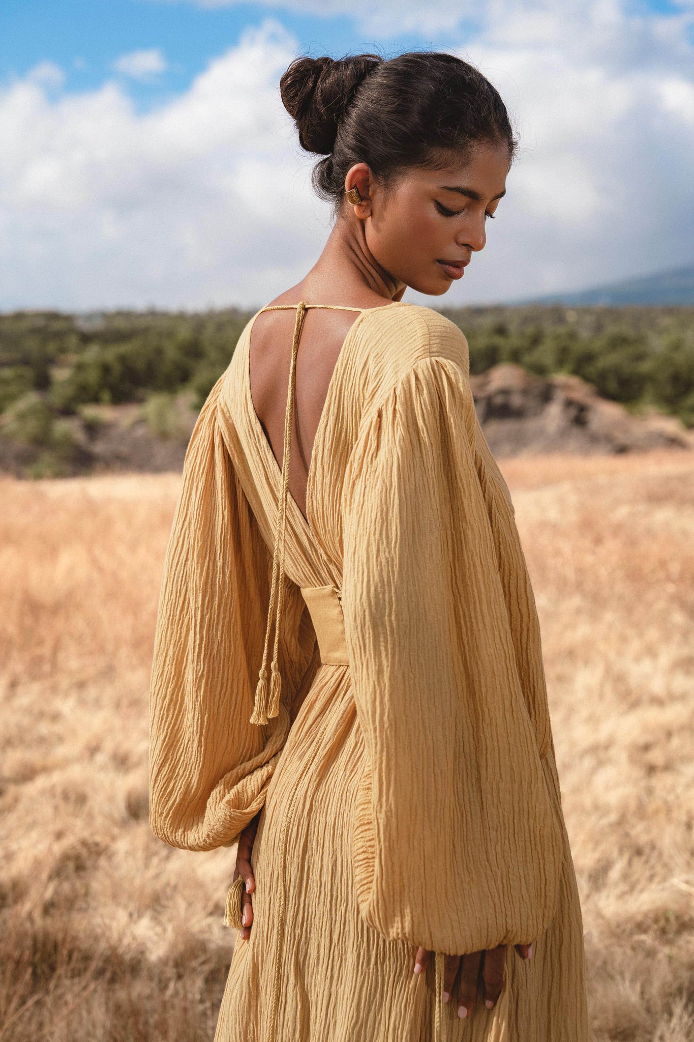 A woman with dark hair in a bun stands in a grassy field, wearing the AYA Sacred Wear Tatva Dress Saffron Gold Colour—an organic, loose-fit dress with long sleeves and deep V-back—framed by trees and a partly cloudy sky.