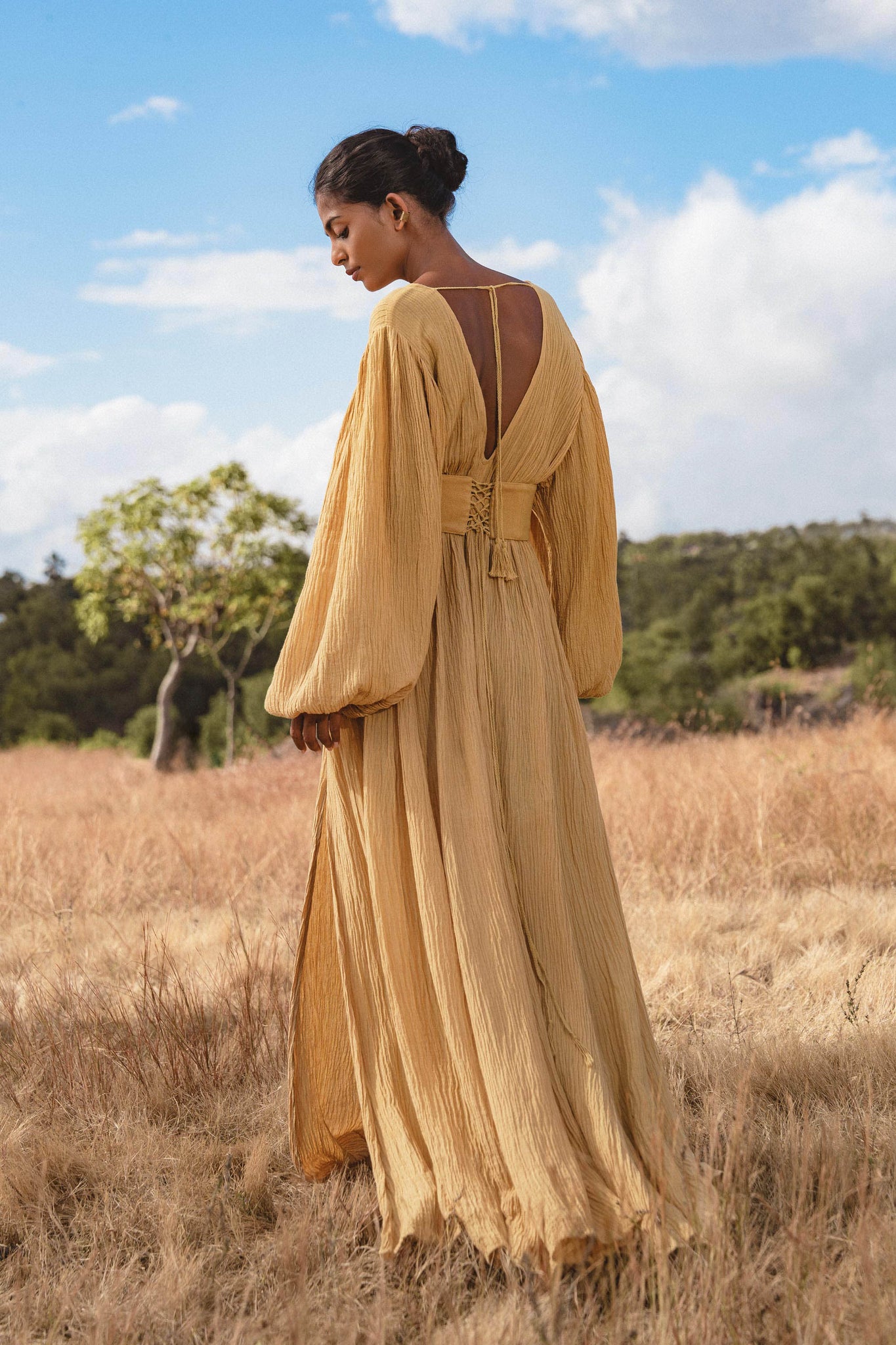 In a dry grassy field, a woman wearing the Tatva Dress Saffron Gold Colour by AYA Sacred Wear stands beneath a partly cloudy sky. The organic, flowing dress features billowy sleeves and an open back, her hair tied as trees frame the serene scene.