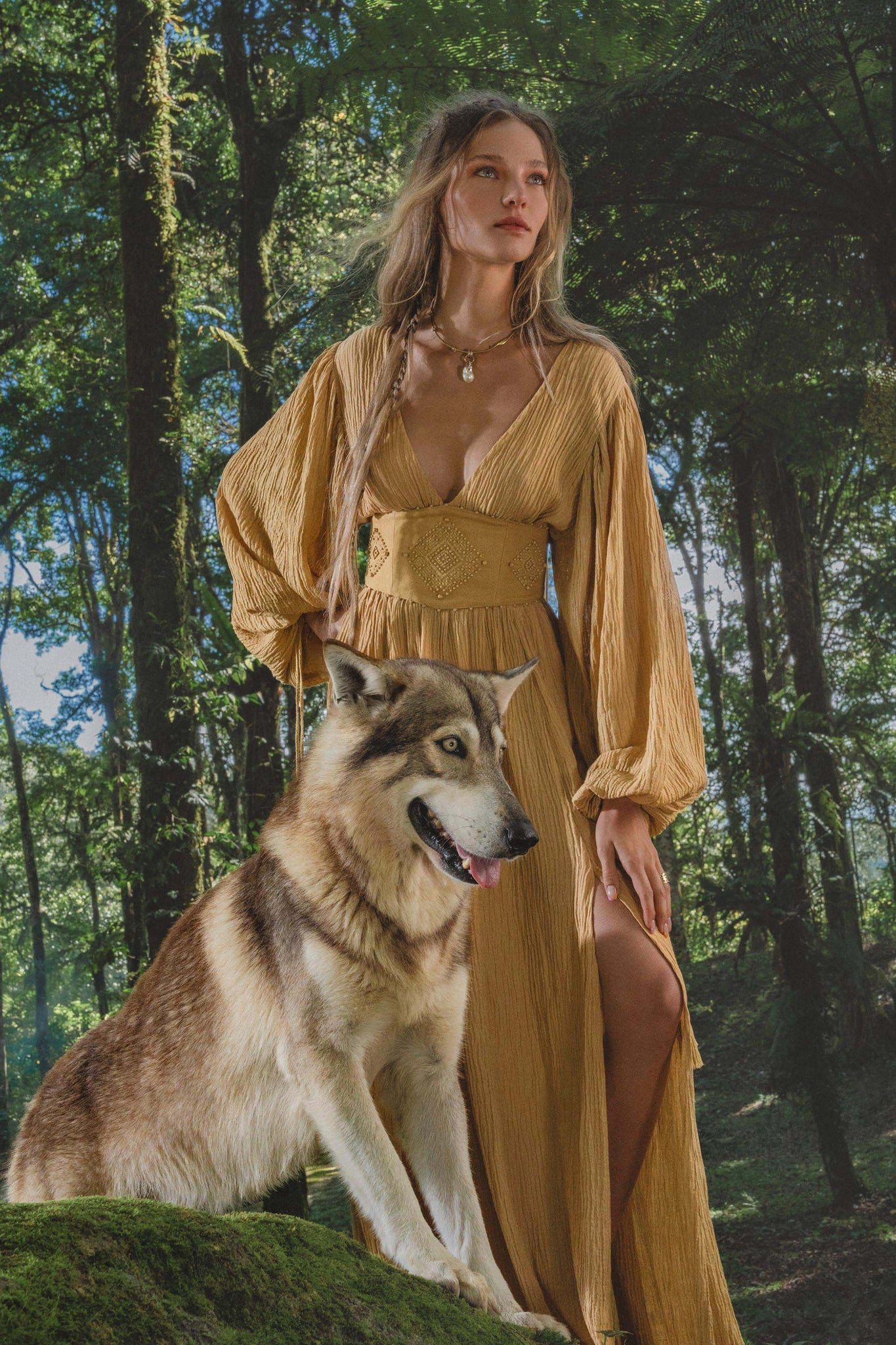 A woman with long wavy hair wears the Tatva Dress Saffron Gold Colour by AYA Sacred Wear, standing calmly in a lush forest with sunlight streaming through the trees and her hand resting on a large, wolf-like dog beside her.