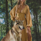 A woman with long wavy hair wears the Tatva Dress Saffron Gold Colour by AYA Sacred Wear, standing calmly in a lush forest with sunlight streaming through the trees and her hand resting on a large, wolf-like dog beside her.