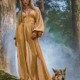 A woman in a flowing Tatva Dress Saffron Gold Colour by AYA Sacred Wear stands barefoot on moss in a misty forest beside a serene wolf-like dog, surrounded by ferns and tall trees with sunlight filtering through the air.