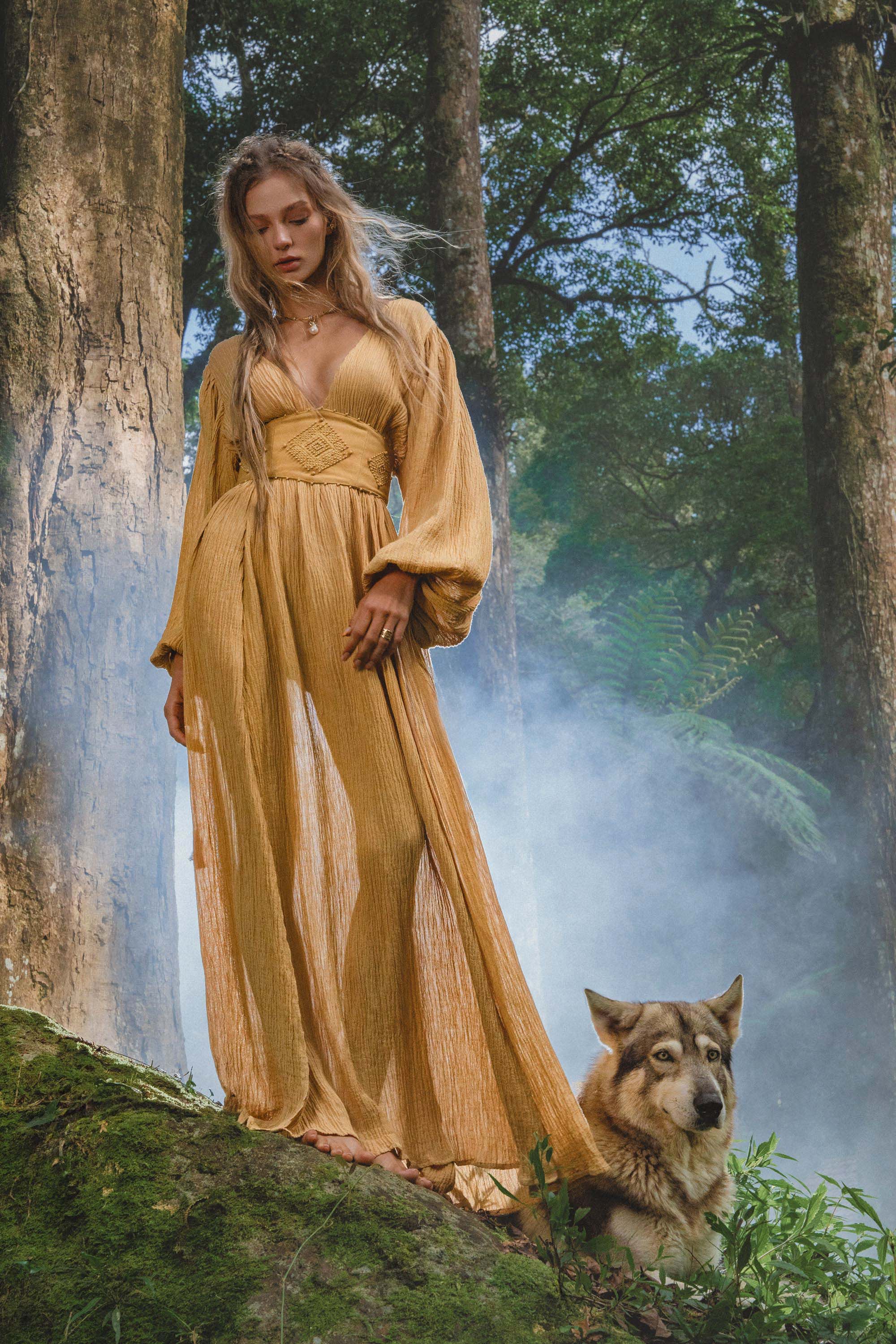 A woman in a flowing Tatva Dress Saffron Gold Colour by AYA Sacred Wear stands barefoot on moss in a misty forest beside a serene wolf-like dog, surrounded by ferns and tall trees with sunlight filtering through the air.