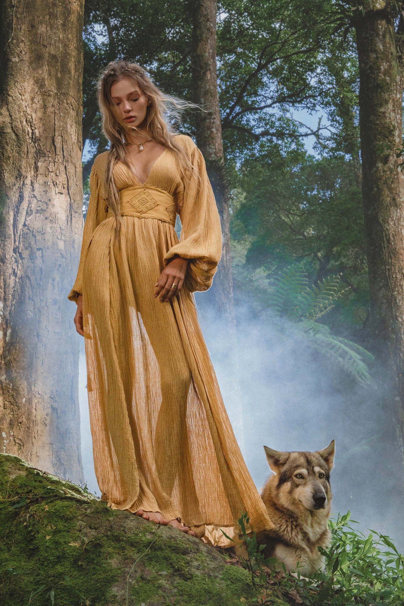 A woman in a flowing Tatva Dress Saffron Gold Colour by AYA Sacred Wear stands barefoot on moss in a misty forest beside a serene wolf-like dog, surrounded by ferns and tall trees with sunlight filtering through the air.