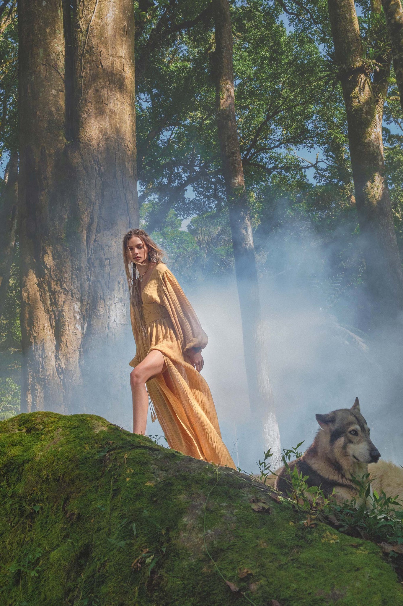 A woman in an AYA Sacred Wear Tatva Dress Saffron Gold Colour – Organic Goddess Dresses stands on a mossy rock in a sunlit forest, mist drifting among the trees as a watchful gray wolf lies beside her under filtered sunlight.