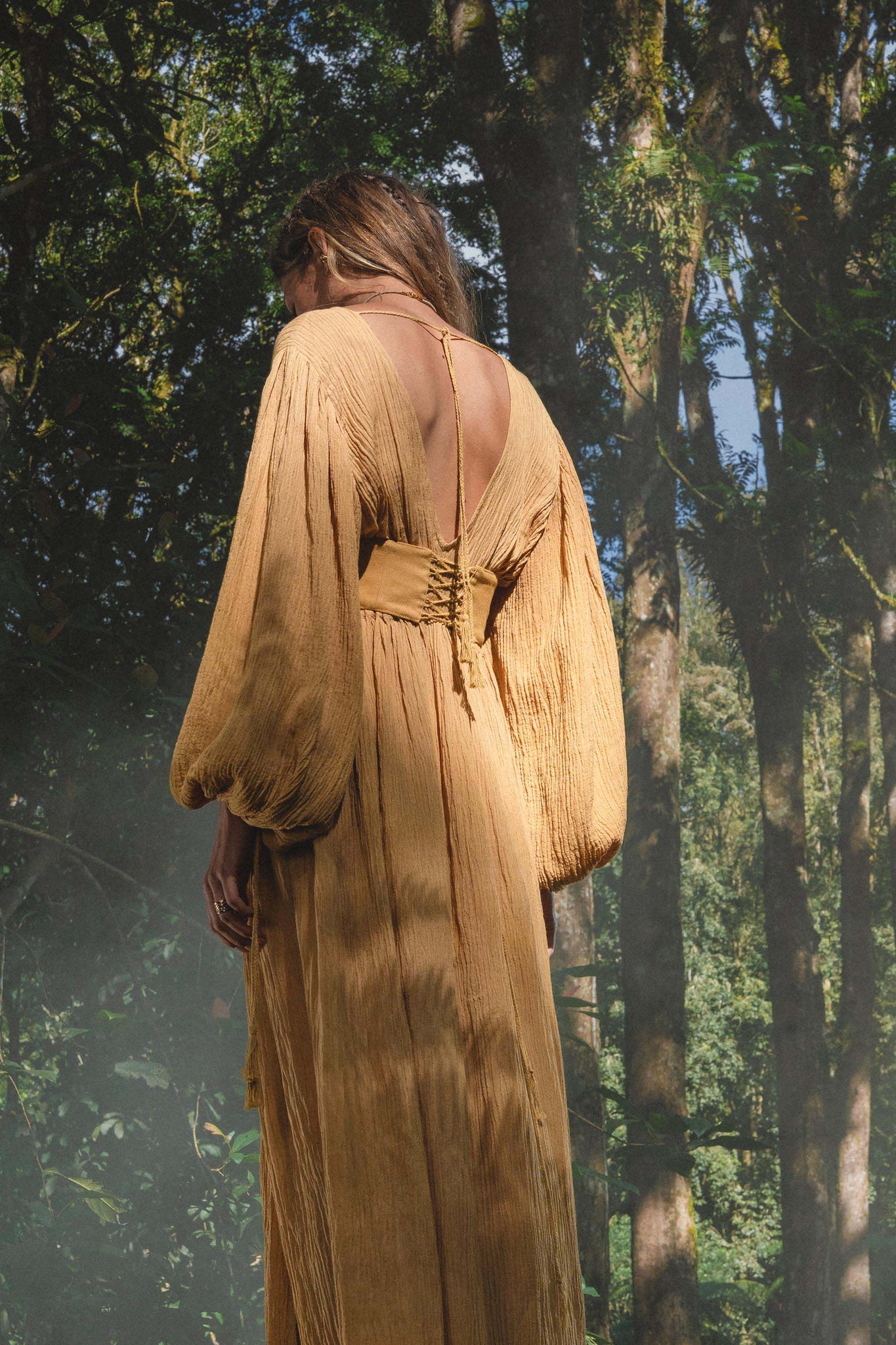 A person stands in a sunlit forest wearing the AYA Sacred Wear Tatva Dress in Saffron Gold—a botanically dyed, organic dress with voluminous sleeves and an open back tied at the neck. Soft light creates a serene, natural setting.