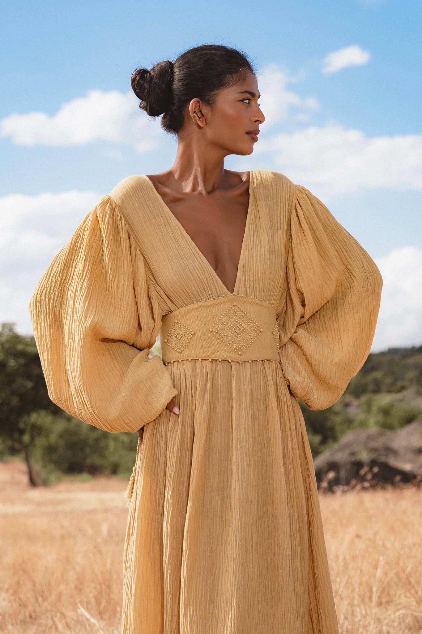 A woman stands in a dry, grassy field wearing the Tatva Dress Saffron Gold Colour by AYA Sacred Wear—a flowing, botanically dyed dress with voluminous long sleeves and deep V-neckline—as she gazes to the side under a blue sky.
