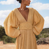A woman stands in a dry, grassy field wearing the Tatva Dress Saffron Gold Colour by AYA Sacred Wear—a flowing, botanically dyed dress with voluminous long sleeves and deep V-neckline—as she gazes to the side under a blue sky.