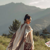 Wearing the "Unique Off-White Boho Poncho with Hand Loomed Tassels" by AYA Sacred Wear, a person gracefully flies a kite within a mountainous landscape. Facing to the right, they are surrounded by sunlit mountains and trees, forming a serene and picturesque scene.