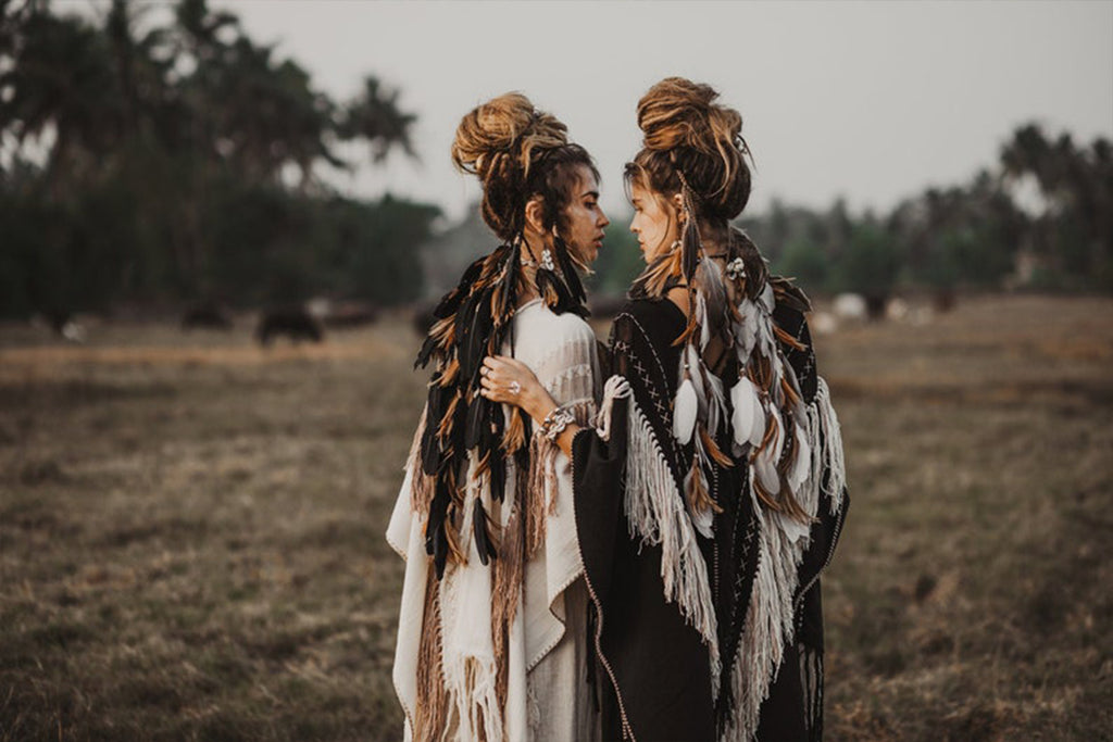 Two people stand in a field, facing each other with arms gently resting on each other's shoulders. They are dressed in bohemian-style clothing, including the Unique Off-White Boho Poncho with Hand Loomed Tassels by AYA Sacred Wear. The background features trees and distant animals under a grey sky.