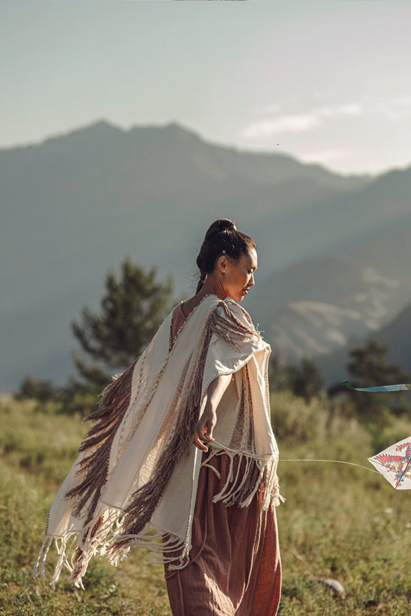 Wearing the "Unique Off-White Boho Poncho with Hand Loomed Tassels" by AYA Sacred Wear, a person gracefully flies a kite within a mountainous landscape. Facing to the right, they are surrounded by sunlit mountains and trees, forming a serene and picturesque scene.