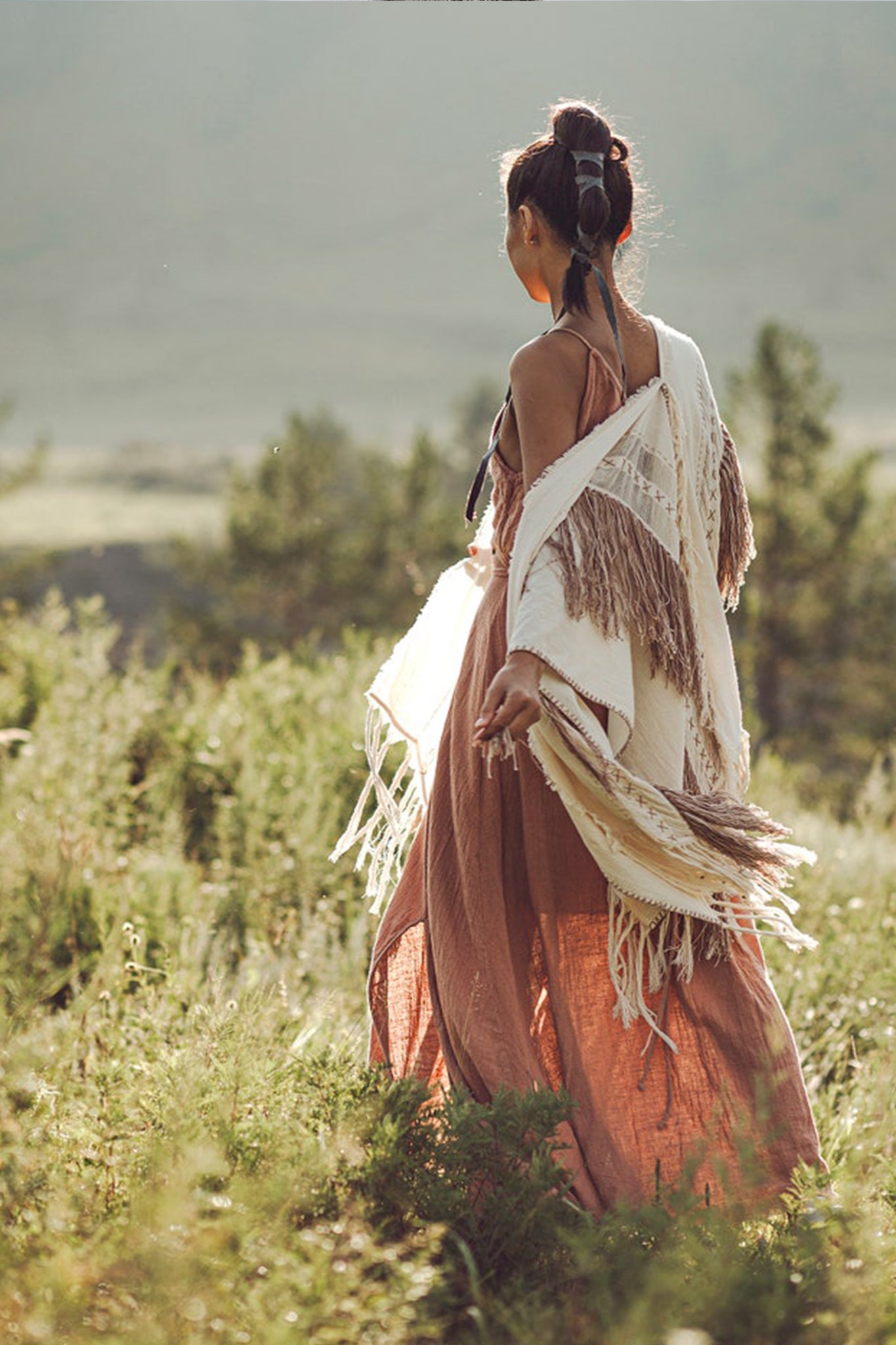 In a lush, sunlit field, a woman stands wearing a flowing pink dress paired with AYA Sacred Wear's Unique Off-White Boho Poncho featuring hand loomed tassels. Her hair is styled in a loose bun adorned with decorative feathers as she gazes into the distance, enveloped by greenery and soft, natural light.