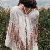 A woman stands outdoors with her back to the camera, draped in the Unique Off-White Boho Poncho with Hand Loomed Tassels from AYA Sacred Wear over a dress. The fringed shawl, adorned with exquisite hand embroidery, complements the tall grass swaying under a cloudy sky, evoking a sense of shamanic tranquility.