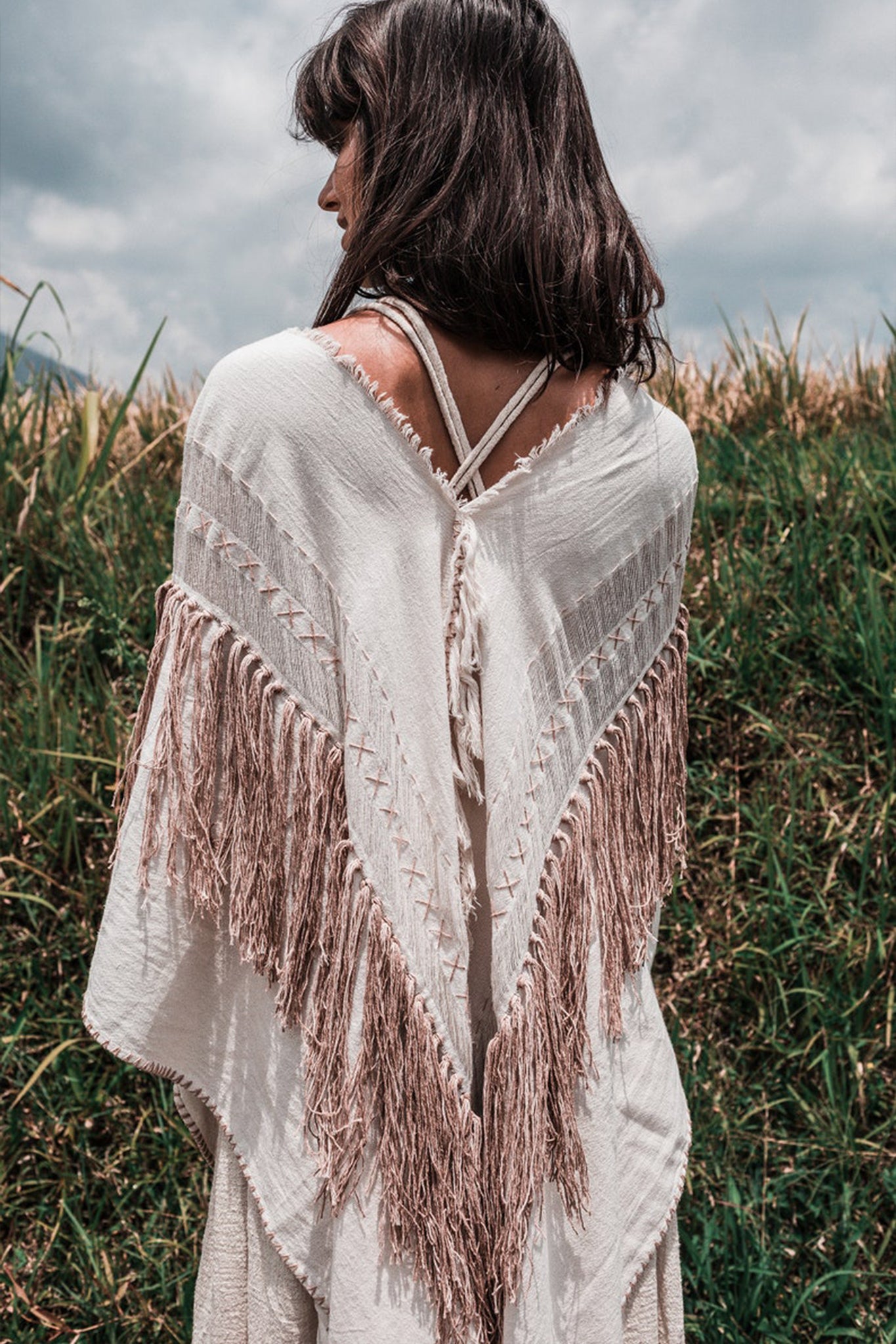 A woman stands outdoors with her back to the camera, draped in the Unique Off-White Boho Poncho with Hand Loomed Tassels from AYA Sacred Wear over a dress. The fringed shawl, adorned with exquisite hand embroidery, complements the tall grass swaying under a cloudy sky, evoking a sense of shamanic tranquility.