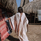 A person wearing the AYA Sacred Wear's Unique Off-White Boho Poncho with Hand Loomed Tassels strolls through a village filled with traditional thatched huts, admiring the hand embroidery on colorful woven textiles. The bright, sunlit scene highlights the intricate artistry and craftsmanship.