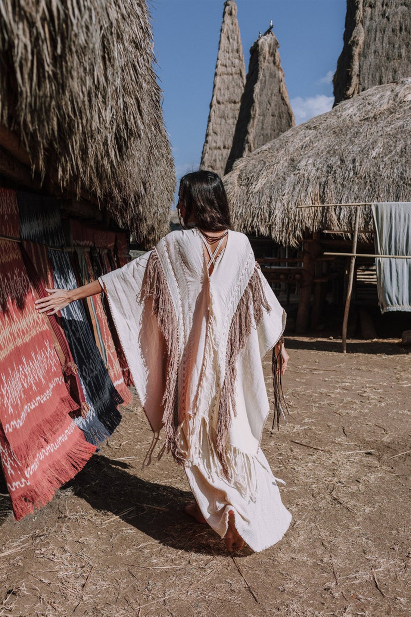 A person wearing the AYA Sacred Wear's Unique Off-White Boho Poncho with Hand Loomed Tassels strolls through a village filled with traditional thatched huts, admiring the hand embroidery on colorful woven textiles. The bright, sunlit scene highlights the intricate artistry and craftsmanship.