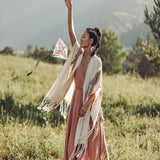 A woman in a long dress and an AYA Sacred Wear Unique Off-White Boho Poncho with Hand Loomed Tassels stands in a grassy field, holding a small kite. Mountains and a clear sky form the serene background, suggesting a peaceful, sunny day.