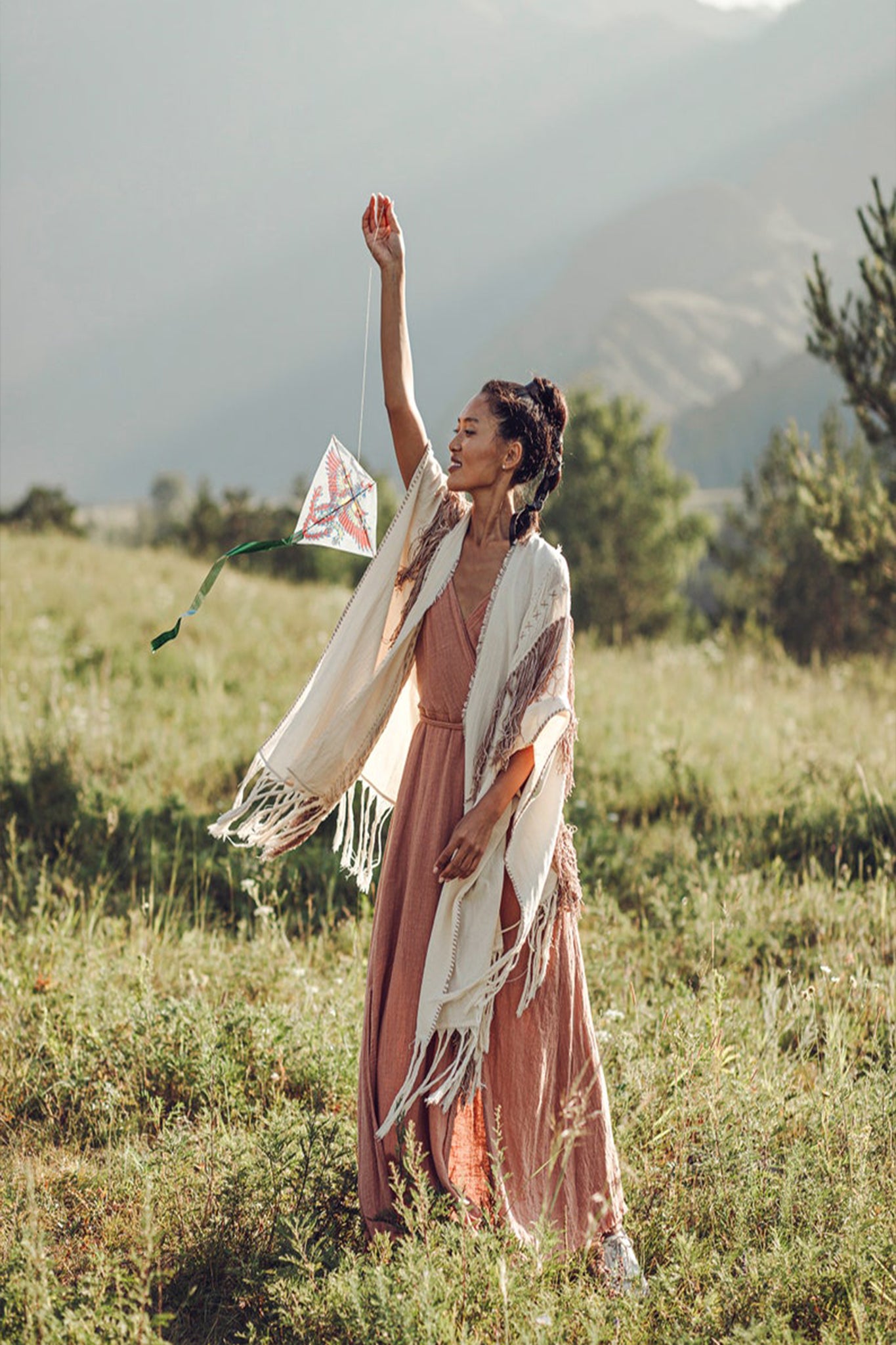 A woman in a long dress and an AYA Sacred Wear Unique Off-White Boho Poncho with Hand Loomed Tassels stands in a grassy field, holding a small kite. Mountains and a clear sky form the serene background, suggesting a peaceful, sunny day.