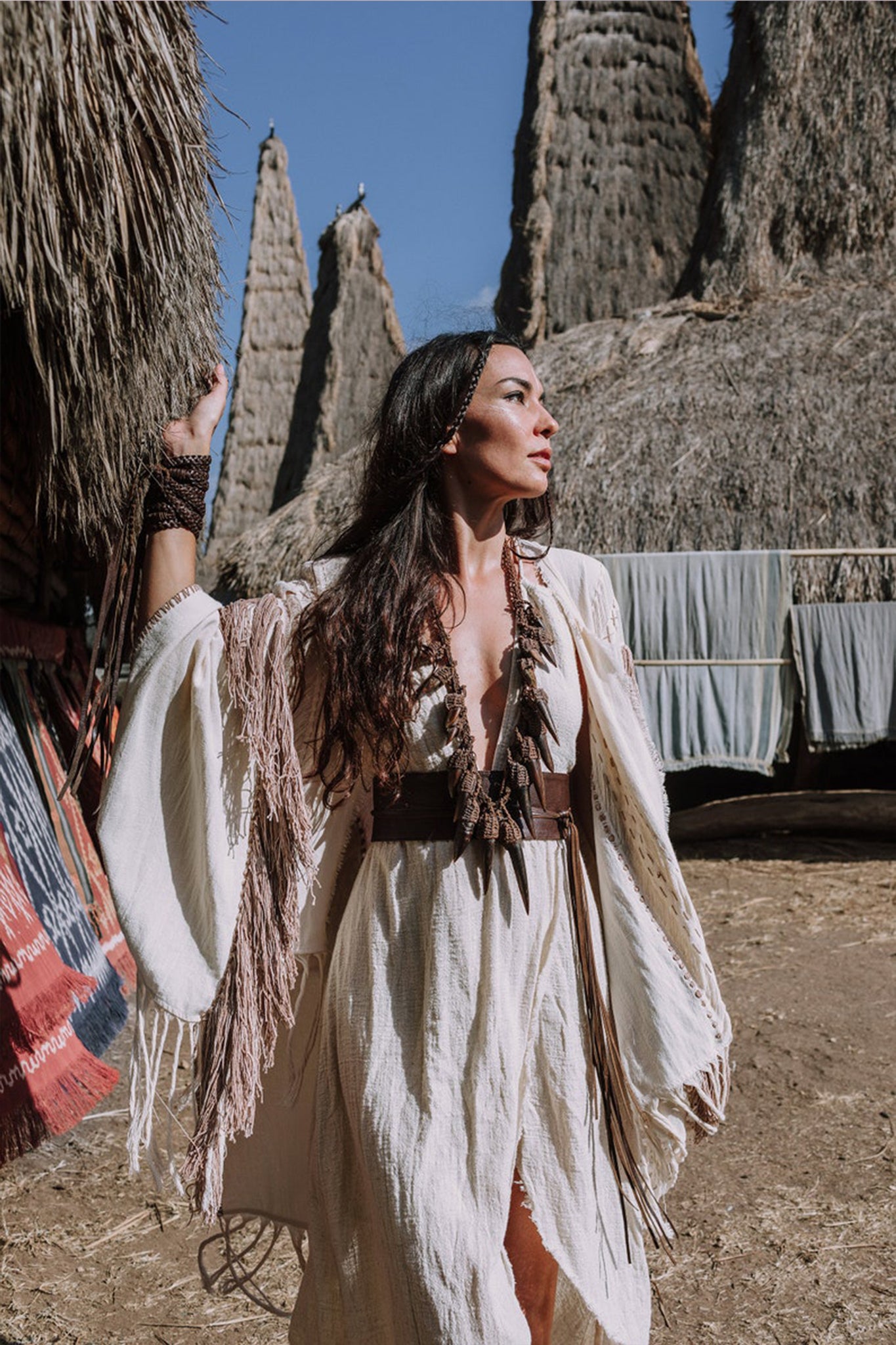 A woman stands outdoors in a Unique Off-White Boho Poncho with Hand Loomed Tassels by AYA Sacred Wear, adorned with bead necklaces. She gazes to the side amidst tall, thatched structures and colorful textiles hanging nearby under a clear blue sky.