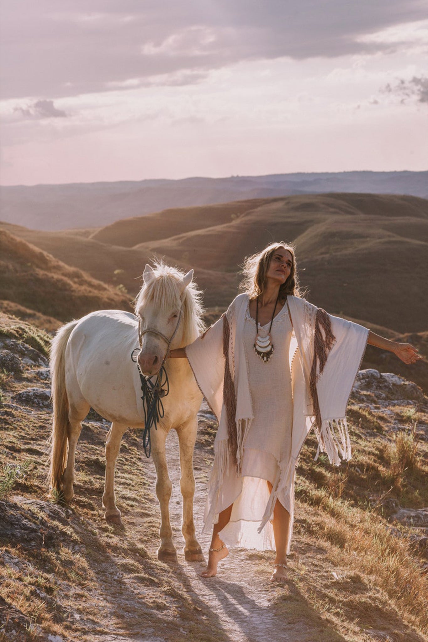 A person stands on a scenic path with a white horse, adorned in an elegant flowing dress layered with AYA Sacred Wear's Unique Off-White Boho Poncho, featuring delicate handloomed tassels and intricate embroidery. The landscape of rolling hills is bathed in warm, golden light, creating a serene and picturesque atmosphere.