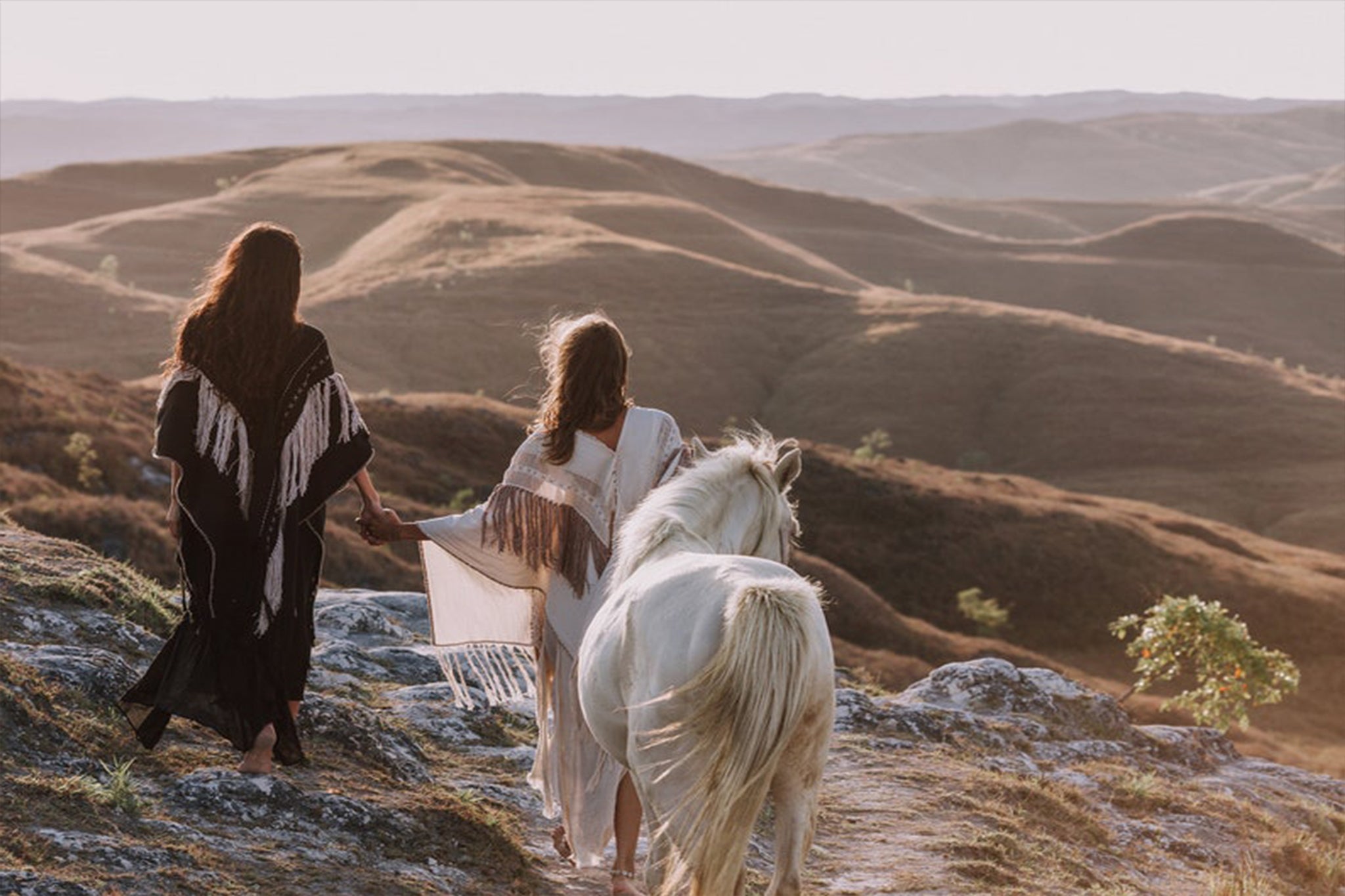 Two people in flowing garments walk hand in hand with a white horse across a rocky hillside at sunset, one of them elegantly draped in AYA Sacred Wear's Unique Off-White Boho Poncho featuring Hand Loomed Tassels. Rolling hills stretch out in the background under a soft, dimming sky.